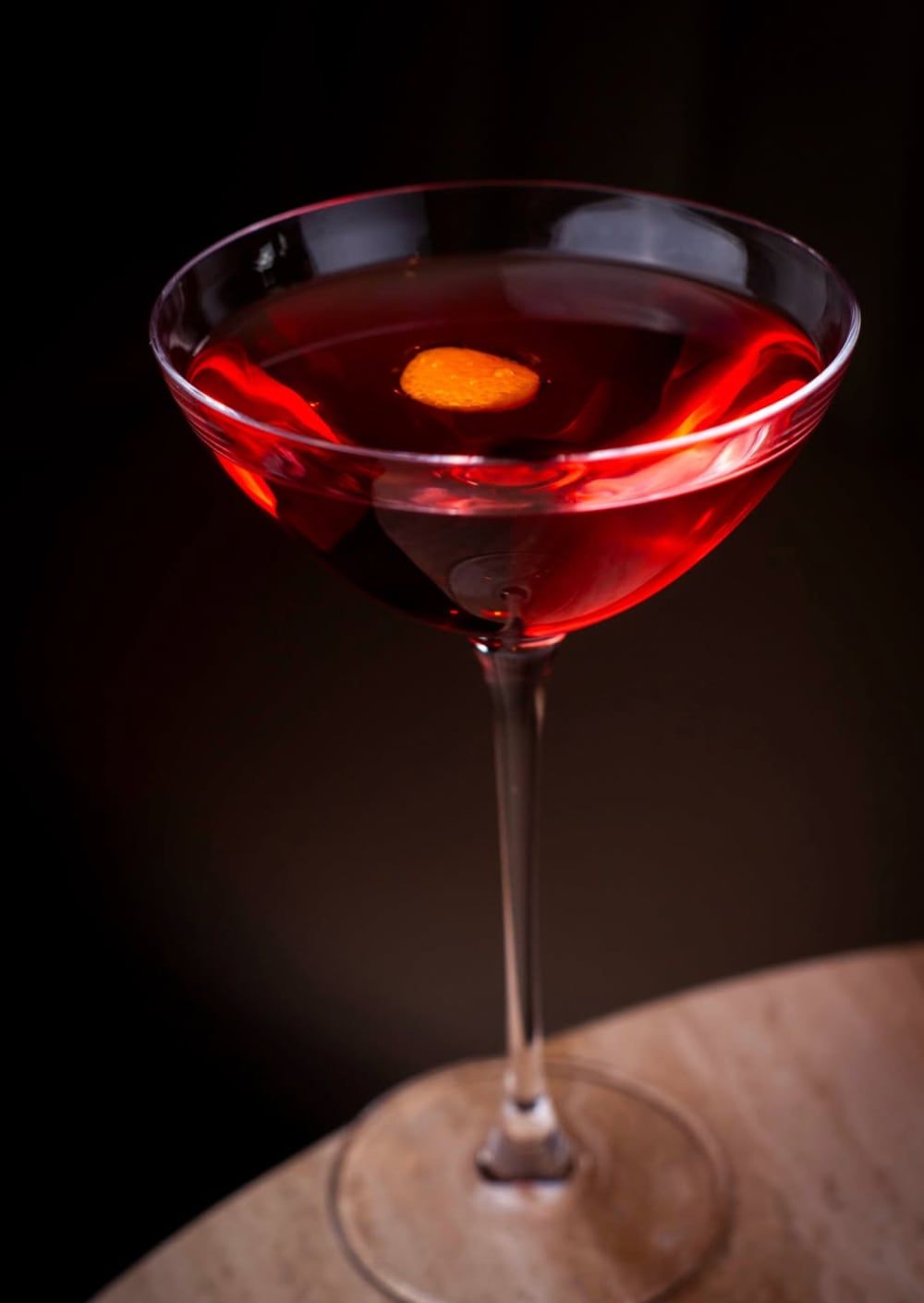 En rød rørt cocktail i en coupe med orangezest, på et lyst stenbord